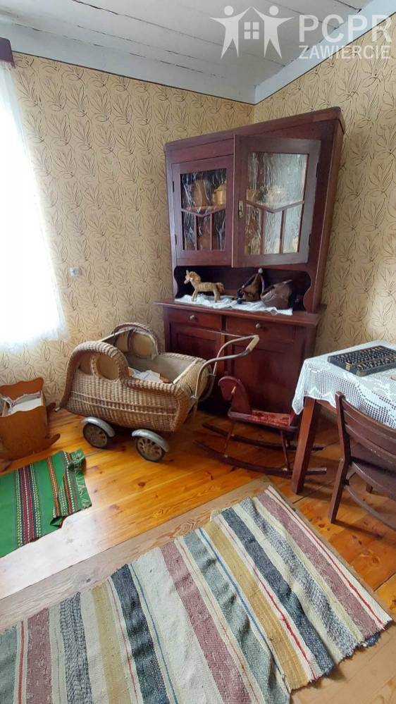 Zdjęcie: Izba w skansenie. Widać regał z eksponatami, garnkami, obok stoi zabytkowa kołyska, stół i konik na biegunach. Na podłodze pleciony dywan.